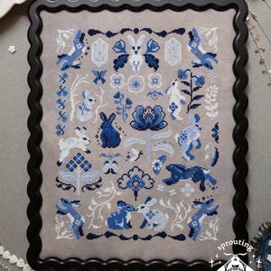 May include: A cross-stitch embroidery on light gray fabric featuring a repeating pattern of rabbits, flowers, and geometric shapes in shades of dark blue, light blue, and white. The piece is framed in a black scalloped frame with the text sprouting lupine.