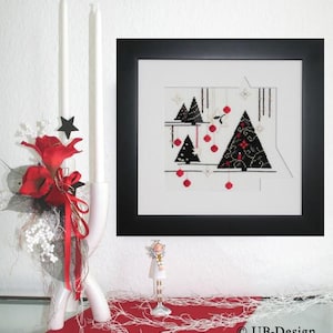 May include: A framed cross-stitch artwork depicting black Christmas trees with red baubles. A white candle holder with two white candles, red flowers, and a decorative angel figurine are also present. The scene is set on a red surface.