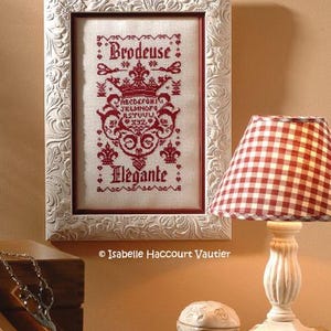 May include: A framed cross-stitch sampler with the words "Brodeuse" and "Élégante" in red thread. The white frame has a floral embossed design. A red and white checkered lamp and a small wooden table are in the foreground.
