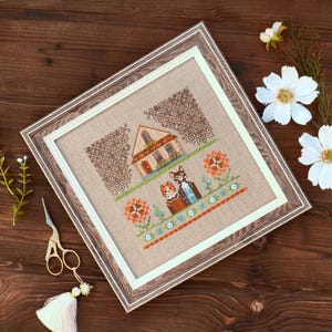 Peut inclure: Image d'une broderie au point de croix encadrée, représentant une maison et deux chats. Le motif comprend une maison, des fleurs et deux chats, avec une palette de couleurs marron, orange et bleu. Le cadre est en bois.