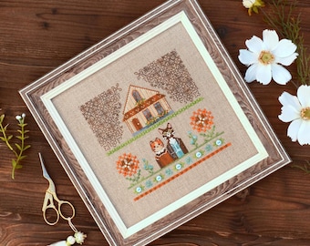 May include: Framed cross-stitch artwork featuring a house and two cats. The artwork is in a wooden frame with a white mat. The cross-stitch design includes a house, flowers, and a pair of cats. The color palette includes brown, orange, and blue.