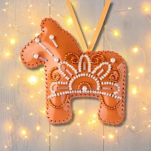 May include: A handcrafted orange Dala horse ornament with intricate beadwork. The horse features white, gold, and clear beads, with a stitched outline. A gold ribbon is attached for hanging. This festive ornament is perfect for holiday decor.