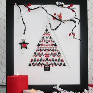May include: Framed cross-stitch Christmas tree with red, black, and white details. The artwork is framed in a black frame and decorated with a star ornament and a garland of berries. Two lit candles are in the foreground.