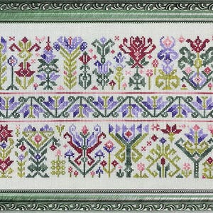Op de afbeelding: Een ingelijste kruissteekborduurwerk met een bloemenontwerp in paars, rood, groen en roze. Het kunstwerk is ingelijst in een groene, sierlijke lijst. Het ontwerp bevat gestileerde bloemen en geometrische patronen.