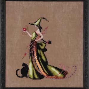 May include: Cross-stitch artwork of a woman in a green witch costume holding a red apple. The witch wears a pointed hat and a long, flowing dress. A black cat sits at her feet. The artwork is framed in a black frame.