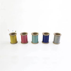 Puede incluir: Cinco carretes de hilo de madera dispuestos en una fila horizontal, con colores amarillo, rosa, azul verdoso, azul y gris. El carrete amarillo del extremo izquierdo contiene tres alfileres de costura con cabezas pequeñas de colores.
