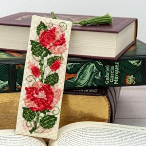 May include: A cross-stitched bookmark with a floral design of red and pink roses and green leaves. The bookmark has a green tassel and is placed in an open book, with other books in the background.