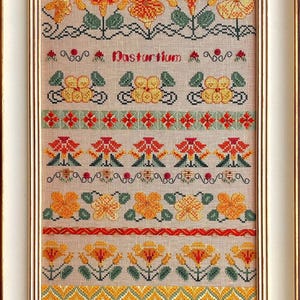 May include: Framed cross-stitch artwork featuring a floral design with orange and yellow flowers, green leaves, and the word "Nasturtium." The artwork is in a white frame with a beige background, showcasing detailed needlework.