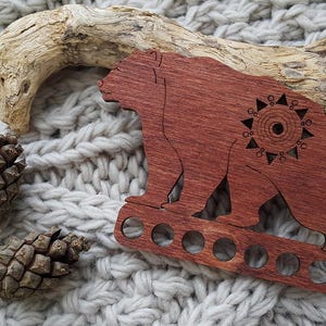 May include: A wooden bear-shaped bottle opener in a reddish-brown hue, featuring a decorative sunburst design. The opener has a series of circular cutouts. Two pine cones and a piece of driftwood are also visible.