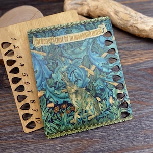 May include: Wooden needle gauge and a needlepoint project cover. The cover features a rabbit in a forest scene with blue and green foliage and the text "The beasts that be in woodland waste". The gauge has numbered holes.