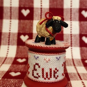 May include: A small carved wooden sheep figurine with a black face and legs, wearing a red string with a gold heart charm, standing on a red wooden spool wrapped in white fabric featuring cross-stitched hearts and the text Ewe.