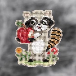 May include: A cross-stitch pattern featuring a cute raccoon holding a red apple. The raccoon is surrounded by green leaves and red flowers.