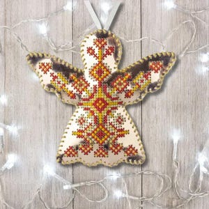 May include: A handmade angel ornament with a white body and wings. The angel features a detailed cross-stitch design in red, yellow, and orange. The wings have a brown border. A white ribbon is attached for hanging. The ornament is approximately 4 inches tall.