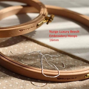 Op de afbeelding: Twee houten borduurringen, de ene bovenop de andere, met een witte draad en een naald. De ringen zijn geëtiketteerd met "Nurge No. 5" en "Nurge No. 1". De tekst "Nurge Luxury Beech Embroidery Hoops 16mm" is zichtbaar in de afbeelding.