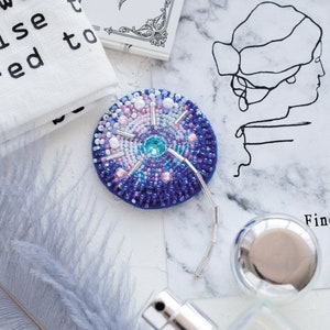 May include: A blue and white beaded brooch with a silver chain attached. The brooch is round and has a sparkly blue center. The brooch is sitting on a white marble surface.
