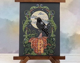 Könnte beinhalten: Kreuzstich-Kunstwerk mit einer schwarzen Krähe auf einem orangefarbenen Kürbis, vor einem Vollmond und einem dunkelgrauen Hintergrund. Das Design umfasst grüne Ranken und Blätter und schafft ein saisonales Halloween-Thema. Das Kunstwerk wird auf einer Holzstaffelei ausgestellt.