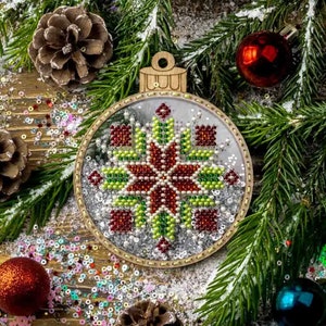 May include: A round, wooden Christmas ornament with a beaded snowflake design in red, green, and white. The ornament is surrounded by pine branches, pine cones, and colorful sequins.