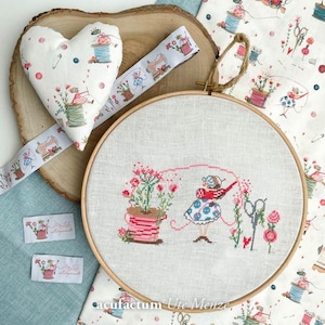 Op de afbeelding: Een hartvormig speldenkussen, lint en borduurring met een bloemig thema. De borduurring heeft een kruissteekontwerp van een meisje met bloemen en een schaar. Het lint en het speldenkussen hebben een bijpassend patroon.