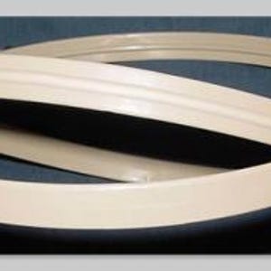 May include: Off-white, curved plastic trim pieces, possibly for furniture or crafts, are arranged in a looping pattern against a dark blue background. The trim has a subtle, raised center detail, and a smooth, matte finish.