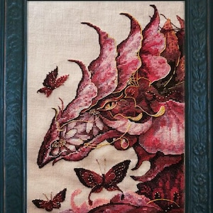 May include: Close-up of a cross-stitch embroidery depicting a pink dragon with gold accents and butterflies. The dragon's eye is visible and the embroidery is framed.