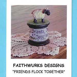 Puede incluir: Un carrete de madera decorativo con una pequeña figura de oveja en la parte superior. El carrete está envuelto en tela con el texto "Friends Flock Together" impreso. El objeto se muestra sobre un tapete de ganchillo blanco. El texto "FAITHWURKS DESIGNS" y "FRIENDS FLOCK TOGETHER" aparece debajo de la imagen.