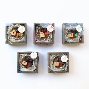 May include: Five gift boxes with clear lids, each containing a small wooden item, a tassel, and a bottle. The boxes are filled with white shredded paper and tied with colorful string. Each box has a tag that says "Cohana".