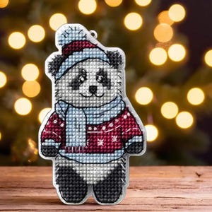 May include: A white panda bear wearing a red and blue sweater and a blue scarf. The panda is wearing a white hat with a red pom-pom. The panda is a Christmas ornament.