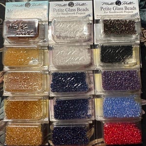 May include: A collection of Mill Hill Petite Glass Beads in various colors, packaged for needlework projects. The beads are displayed in clear plastic containers, showcasing shades like clear, black, blue, and red. Each package contains 1.60 grams of beads.