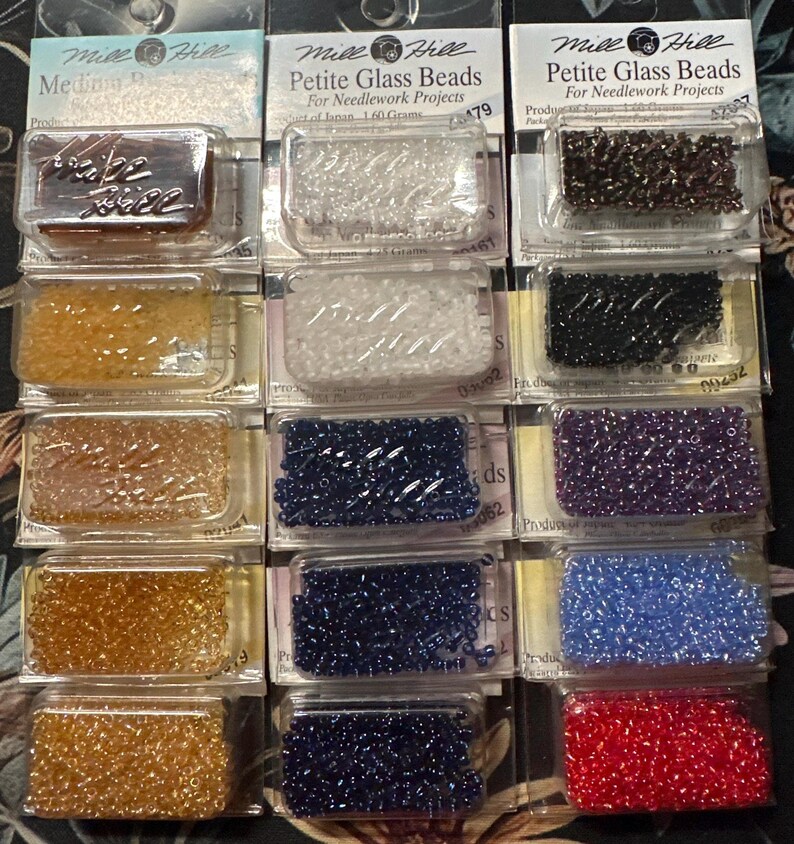 May include: A collection of Mill Hill Petite Glass Beads in various colors, packaged for needlework projects. The beads are displayed in clear plastic containers, showcasing shades like clear, black, blue, and red. Each package contains 1.60 grams of beads.