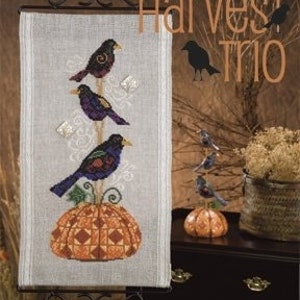 Könnte beinhalten: Ein Kreuzstichmuster mit drei lila und schwarzen Vögeln, die auf einem Ast über einem Kürbis sitzen. Das Muster trägt den Titel "Harvest Trio" von Jim Shore.