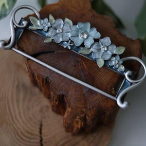 May include: A silver-toned barrette featuring a row of light blue floral embellishments with crystal centers and green leaves. The barrette has a curved design with decorative swirls on each end. The barrette is set against a textured wooden surface.