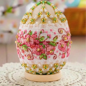 May include: An egg-shaped ornament featuring a central band of white fabric with pink and green floral cross-stitch embroidery. The top and bottom sections are decorated with a lattice pattern of white pearls and metallic green seed beads.