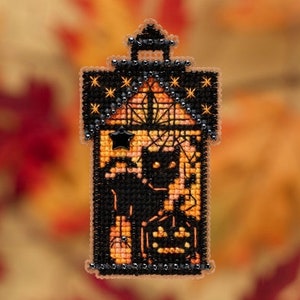 Puede incluir: Un adorno de punto de cruz negro y naranja que representa una casa encantada con un gato negro y calabazas. La casa tiene un techo negro y una estrella blanca en la fachada.