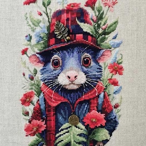 Puede incluir: Imagen de punto de cruz de un roedor azul con sombrero y chaqueta a cuadros rojos, rodeado de flores rojas y rosas y follaje verde. Un dije dorado con forma de hoja y árbol cuelga de la chaqueta. Sobre fondo de tela beige claro.