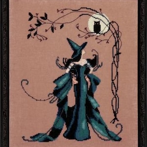 May include: Cross-stitch artwork of a witch in a teal gown and hat, standing beneath a tree with an owl perched on a full moon. The artwork is framed in a black frame, set against a muted pink background.