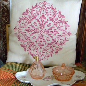 May include: A decorative pillow with a pink floral design rests on a chair. A perfume bottle and a small jar sit on a white tray with a floral pattern. The chair has a woven seat with red, green, and yellow stripes.