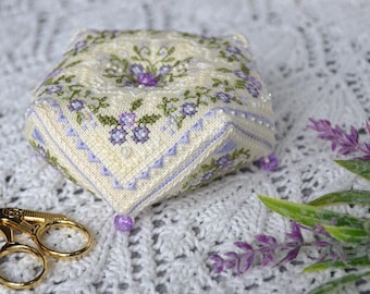 May include: A square, cream-colored pincushion with a floral design in shades of purple and green. The pincushion has a beaded edge and a central button. Gold-colored scissors are next to the pincushion, and sprigs of lavender are in the background.