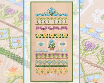 May include: A cross-stitch pattern featuring floral designs in shades of blue, yellow, purple, and green. The design includes various floral motifs, geometric patterns, and a central basket of flowers. The background is a tan fabric.