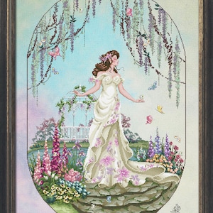 May include: Cross-stitch artwork of a woman in a white gown with pink floral accents, standing in a garden. The scene includes a pond, flowers, butterflies, and a gazebo, all within an oval frame. The artwork is framed in a dark, ornate wooden frame.