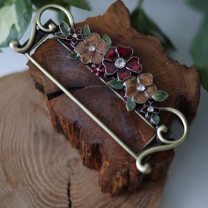 May include: A decorative floral brooch with a gold-tone frame. The brooch features brown and red enameled flowers with crystal accents and green leaves. The brooch is set on a piece of wood.