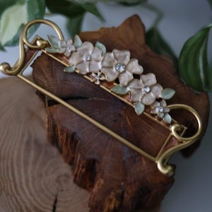 May include: A gold-coloured barrette with a floral design. The barrette has a rectangular frame with curved edges and is decorated with cream-coloured flowers with rhinestone centres and green leaves. The barrette is displayed on a piece of wood.
