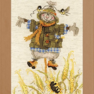 May include: A framed cross-stitch embroidery featuring a scarecrow wearing a green plaid jacket, orange scarf, and blue checkered shirt. The scarecrow has two small birds perched on its hat and stands in a field of yellow corn and sunflowers.