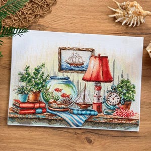 May include: A cross-stitch embroidery piece depicting a nautical-themed shelf with a fishbowl containing two goldfish, a lighthouse-shaped lamp with a red shade, a framed ship painting, a small sailboat model, a clock, books, and potted plants.