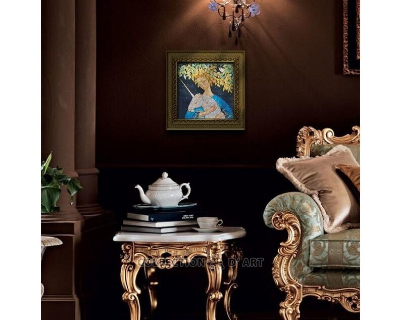 May include: A framed artwork depicts a woman with a unicorn, set against a backdrop of foliage. The artwork is displayed in a room with ornate furniture, including a gold-trimmed side table with a white teapot and books. The room has a dark brown wall.