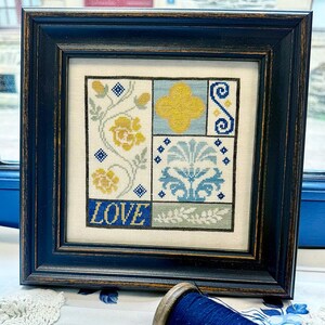May include: Framed cross-stitch artwork with a navy blue frame. The design features floral and geometric patterns in yellow, blue, and gray, with the word "LOVE" stitched in blue at the bottom. The artwork is displayed on a white background.