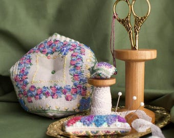 May include: A still life of sewing notions on a gold tray. Includes a floral embroidered pincushion, a small spool of thread, a pair of gold scissors, and a rectangular pincushion with a rainbow design. White lace and pins are also present.