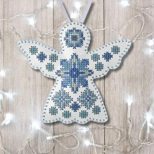 Puede incluir: Adorno blanco en forma de ángel con bordado de punto de cruz azul. Las alas y el cuerpo presentan patrones florales y geométricos. El adorno tiene un borde blanco cosido y un pequeño lazo para colgar. Luces decorativas en el fondo.