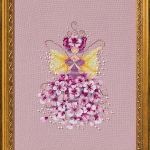May include: Cross-stitch artwork of a fairy in a gold frame. The fairy has a floral dress in shades of pink and white, yellow wings, and a floral crown. The background is a light pink fabric.