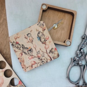 Puede incluir: Una caja de costura de madera con tapa con bisagras, con un estampado caprichoso de gatitos en ramas. La caja está abierta, revelando un imperdible y agujas. También hay tijeras decorativas con mangos florales.