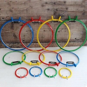 May include: A collection of colorful embroidery hoops in various sizes. The hoops are in blue, red, yellow, and green. The hoops are made of plastic and have a metal clasp. The background is a wooden surface.